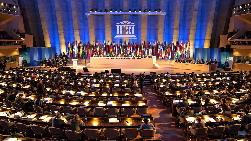 Delegates seated in the UNESCO plenary hall during a conference, with the UNESCO emblem and national flags on stage.